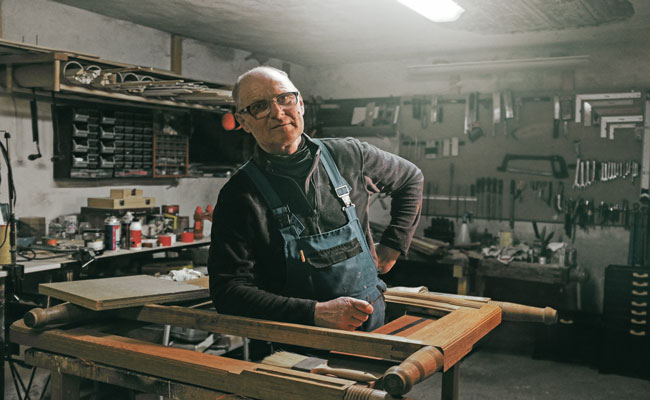 Ein Mann in Latzhose lehnt in einer Werkstatt an der Werkbank. 