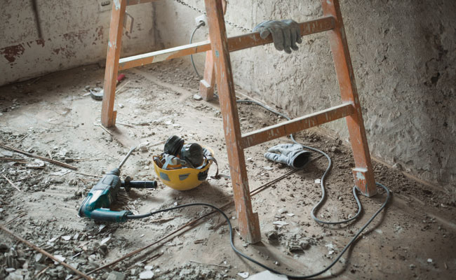 Auf einem dreckigen Baustellenboden liegen eine Bohrmaschine, ein Sicherheitshelm mit Gehörschutz und Handschuhe. Eine Leiter steht daneben.